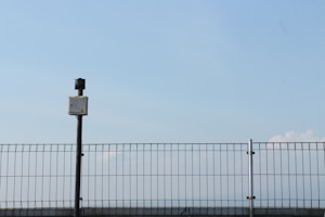 A metal fence under a clear blue sky with a surveillance camera positioned on top of a black pole. The pole and camera are centralized, with the fence extending across the width of the image. The sky is vast and mostly clear with a few faint clouds visible on the right side.