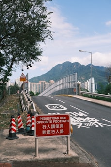 A road with a metal barrier and a prominent red sign instructing pedestrians to use the opposite footpath. The surrounding area includes trees and a view of distant mountains under a clear blue sky. Traffic cones and a speed limit marking of 30 are visible on the road.