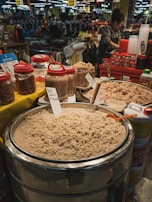 A bustling market scene featuring large containers filled with what appears to be fermented seafood surrounded by various jars with red lids. A vendor, wearing a striped shirt and apron, stands nearby. In the background, shelves are filled with merchandise and there are people shopping.