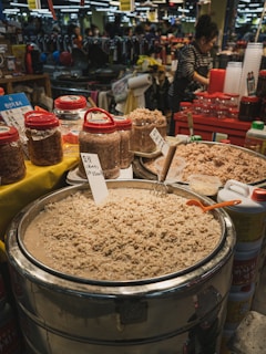 A bustling market scene featuring large containers filled with what appears to be fermented seafood surrounded by various jars with red lids. A vendor, wearing a striped shirt and apron, stands nearby. In the background, shelves are filled with merchandise and there are people shopping.