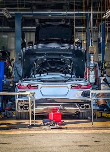 A silver sports car with its trunk open is stationed inside a garage or workshop, with various tools and equipment scattered around. The car is positioned on a lift, and there is a red fuel container on the ground behind it.