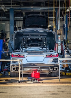 A silver sports car with its trunk open is stationed inside a garage or workshop, with various tools and equipment scattered around. The car is positioned on a lift, and there is a red fuel container on the ground behind it.