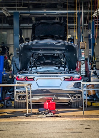 A silver sports car with its trunk open is stationed inside a garage or workshop, with various tools and equipment scattered around. The car is positioned on a lift, and there is a red fuel container on the ground behind it.
