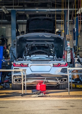 A silver sports car with its trunk open is stationed inside a garage or workshop, with various tools and equipment scattered around. The car is positioned on a lift, and there is a red fuel container on the ground behind it.