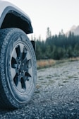 A close-up view of a rugged vehicle tire on a gravel road, with a forest and mountains in the background. The tire is marked with the brand Goodyear and features a multi-ribbed tread pattern suggesting off-road capabilities. The background shows a blend of green trees and a distant, slightly blurred mountain range under a clear sky.
