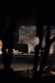 A dark, shadowy foreground frames an abandoned, partially constructed or ruined building. Smoke or mist drifts across the scene, adding a mysterious atmosphere, while leafless tree branches are visible through an open section of the structure.