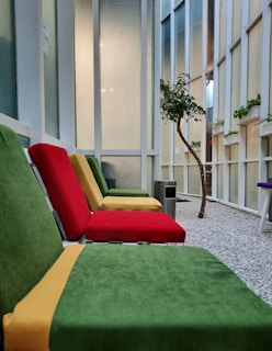 The image features a seating area with colorful cushioned chairs in green, red, and yellow. The setting appears to be indoors with a modern design, featuring tall windows and a gravel floor. A small tree and several potted plants add a touch of nature to the space. There is a metal trash can nearby, contributing to a clean and organized atmosphere.