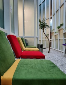 The image features a seating area with colorful cushioned chairs in green, red, and yellow. The setting appears to be indoors with a modern design, featuring tall windows and a gravel floor. A small tree and several potted plants add a touch of nature to the space. There is a metal trash can nearby, contributing to a clean and organized atmosphere.