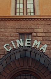 An ornate, vintage-style cinema entrance featuring large, bold letters spelling 'CINEMA' above an arched doorway. The facade is built with textured stonework and a pair of tall, narrow windows is visible above.