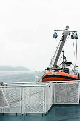 A lifeboat is secured on the deck of a ship. The deck is surrounded by white metal railings, and the background shows a misty sea with an overcast sky. An island can be seen in the distance.