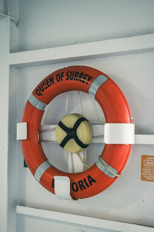 A bright orange life preserver with the words 'QUEEN OF SURREY' and 'VICTORIA' is mounted on a white wall. The device is secured with white straps and has a rope coiled in its center. A small warning sign is visible nearby.