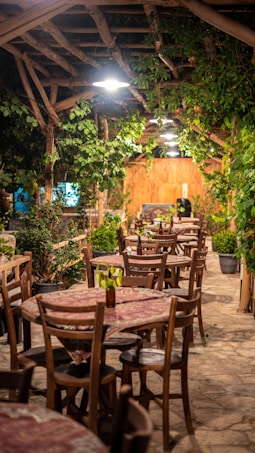 A rustic outdoor dining area with multiple wooden tables and chairs. The tables are adorned with patterned tablecloths, and the space is surrounded by lush green plants and wood beams overhead. Soft lighting enhances the cozy atmosphere, and potted plants are placed throughout the area.