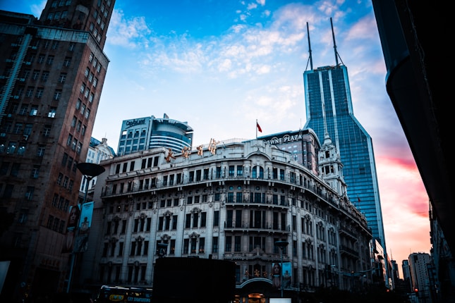 A bustling urban scene featuring historic and modern architecture, highlighted by a colorful sky during sunset. The buildings are tall and imposing, with signs and advertisements visible on the facades.