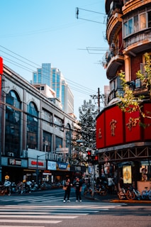 A bustling urban street corner featuring a mix of modern and traditional architecture. The street is lined with various shops and a multitude of bicycles, indicating a busy city environment. Two people stand on a pedestrian crossing, while others can be seen in the background going about their day. Vibrant red signage with Chinese characters is prominently displayed on one of the buildings.