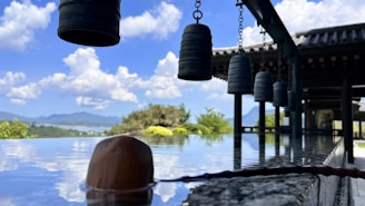 A tranquil outdoor scene features a series of hanging metal bells under a traditional Asian structure with a tiled roof. The reflection of the sky and clouds is visible on a smooth reflective surface, possibly water, with a wooden dipper resting on it. In the background, lush greenery and a distant view of mountains are present.