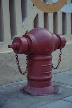 A red fire hydrant with some scratch marks stands on a concrete sidewalk. It is equipped with metal chains and has some text engraved on its surface. The background features a light beige wall with vertical cut-out patterns and a circular design painted partially visible.