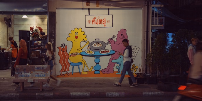 A vibrant street scene featuring a mural on a restaurant wall depicting colorful cartoon characters enjoying a meal at a table. Passersby walk along the sidewalk, and the surrounding area includes potted plants and an open shop with people inside.
