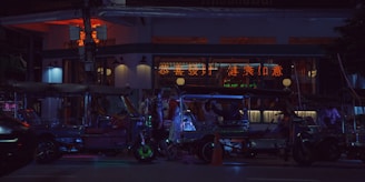 A nighttime street scene featuring several tuk-tuks parked in front of a building with illuminated neon signs in a foreign script. The light from the signs casts a colorful glow on the vehicles and the surrounding area, creating a vibrant atmosphere. A few people are visible near the tuk-tuks, engaging in conversation.
