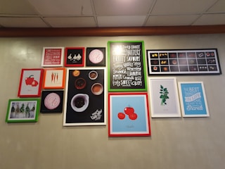 A wall features a collection of framed pictures and artworks, including illustrations of vegetables such as tomatoes and carrots, and images of herbs. Some frames contain typography prints with phrases like 'FRUITY', 'SMOOTH', and 'THE BEST THINGS IN LIFE ARE MEANT TO BE Shared'. The frames vary in color, including red, green, and white borders. The background is a plain, muted color.