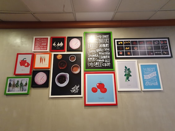 A wall features a collection of framed pictures and artworks, including illustrations of vegetables such as tomatoes and carrots, and images of herbs. Some frames contain typography prints with phrases like 'FRUITY', 'SMOOTH', and 'THE BEST THINGS IN LIFE ARE MEANT TO BE Shared'. The frames vary in color, including red, green, and white borders. The background is a plain, muted color.