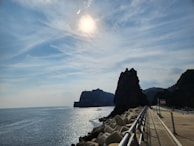 A scenic coastal landscape features a sunlit sky with wispy clouds. The ocean stretches into the distance, bordered by a rocky shoreline and a prominent dark rock formation. A pathway with railings runs beside the ocean, lined with large tetrapod structures for coastal protection.