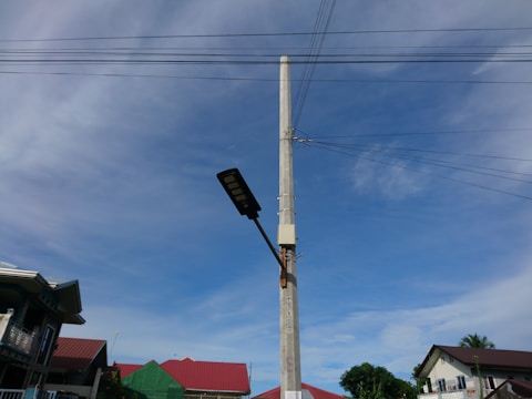 A tall concrete utility pole stands prominently with electrical wires extending from it in different directions. Attached to the pole is a street light. In the background, there are several houses with red roofs against a clear blue sky with some light clouds.