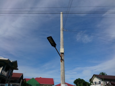 A tall concrete utility pole stands prominently with electrical wires extending from it in different directions. Attached to the pole is a street light. In the background, there are several houses with red roofs against a clear blue sky with some light clouds.