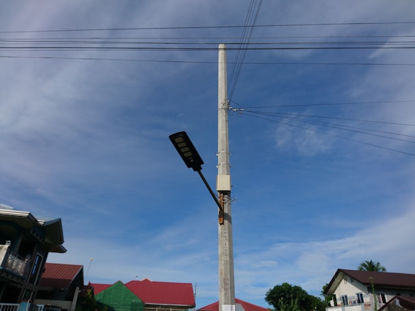 A tall concrete utility pole stands prominently with electrical wires extending from it in different directions. Attached to the pole is a street light. In the background, there are several houses with red roofs against a clear blue sky with some light clouds.