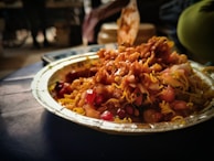 A close-up of a paper plate filled with a colorful and vibrant Indian street food dish, featuring crispy fried snacks, sev, chopped onions, pomegranate seeds, and a drizzle of tamarind sauce. The background is blurred, highlighting the food.