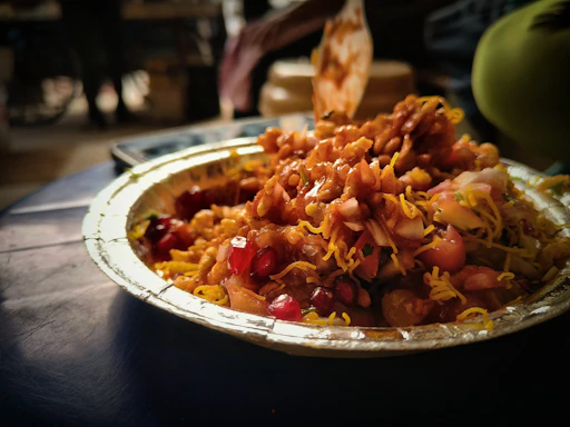 A close-up of a paper plate filled with a colorful and vibrant Indian street food dish, featuring crispy fried snacks, sev, chopped onions, pomegranate seeds, and a drizzle of tamarind sauce. The background is blurred, highlighting the food.