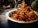 A close-up of a paper plate filled with a colorful and vibrant Indian street food dish, featuring crispy fried snacks, sev, chopped onions, pomegranate seeds, and a drizzle of tamarind sauce. The background is blurred, highlighting the food.
