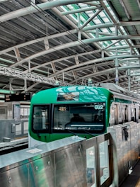 A green train is stationed at a modern platform with a high arched metal roof and several overhead lights. The platform has metal railings and a display board with information. The environment is industrial and urban.