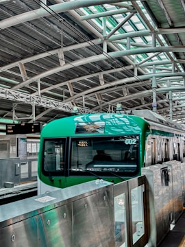 A green train is stationed at a modern platform with a high arched metal roof and several overhead lights. The platform has metal railings and a display board with information. The environment is industrial and urban.