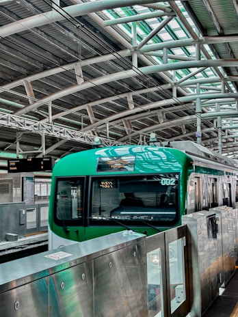 A green train is stationed at a modern platform with a high arched metal roof and several overhead lights. The platform has metal railings and a display board with information. The environment is industrial and urban.