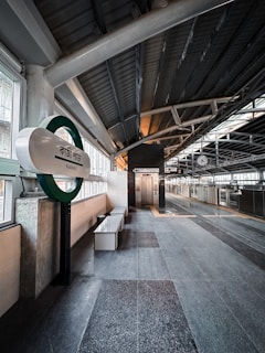 A modern train station platform with an elevated roof structure. The platform appears clean and well-maintained, featuring a sign in another language naming the station as 'Kazipara.' There is a bench along the wall and an elevator in the background. The environment is well-lit with natural light coming from the windows.