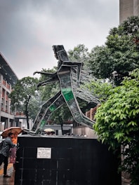 A sculpture made from recycled electronic components depicts a person walking. The sculpture is mounted on a large base with a plaque that reads 'Digital Desire'. Trees and a building are visible in the background, with several people nearby under umbrellas.