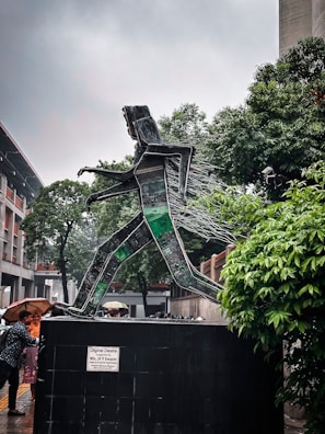 A sculpture made from recycled electronic components depicts a person walking. The sculpture is mounted on a large base with a plaque that reads 'Digital Desire'. Trees and a building are visible in the background, with several people nearby under umbrellas.