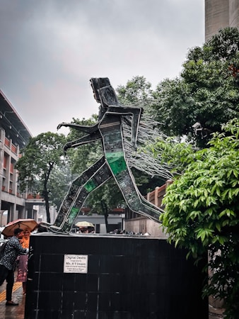 A sculpture made from recycled electronic components depicts a person walking. The sculpture is mounted on a large base with a plaque that reads 'Digital Desire'. Trees and a building are visible in the background, with several people nearby under umbrellas.