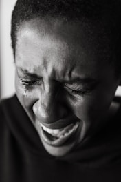 A close-up black and white portrait of a person with short hair, eyes closed, and mouth open as if crying or in emotional distress. Tears are visible on the person's cheek.