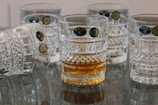 A group of intricately designed glass tumblers with textured patterns, one of which contains a golden liquid, are arranged on a reflective surface. The glasses feature labels indicating their origin from Bohemia.