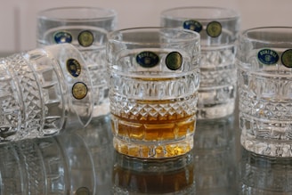 A group of intricately designed glass tumblers with textured patterns, one of which contains a golden liquid, are arranged on a reflective surface. The glasses feature labels indicating their origin from Bohemia.
