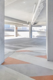 A modern, empty parking garage with a gray and white concrete interior, featuring geometric patterns on the floor with orange and gray colors. Natural light floods the space from large openings, and there are structural pillars supporting the ceiling.