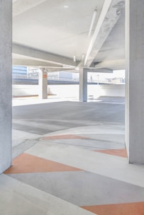 A modern, empty parking garage with a gray and white concrete interior, featuring geometric patterns on the floor with orange and gray colors. Natural light floods the space from large openings, and there are structural pillars supporting the ceiling.