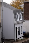 A small, narrow house with gray shingles and white exterior walls is tucked between two larger buildings. The house features two dormer windows on the roof and additional windows on the front facade. The architecture is compact and modest with black trim accentuating the windows and door. The surroundings include a paved sidewalk and autumn foliage in the background.