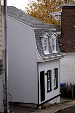 A small, narrow house with gray shingles and white exterior walls is tucked between two larger buildings. The house features two dormer windows on the roof and additional windows on the front facade. The architecture is compact and modest with black trim accentuating the windows and door. The surroundings include a paved sidewalk and autumn foliage in the background.