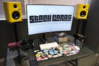 A modern workspace setup featuring a large curved monitor with the words 'STACK CARDS' and 'HUMAN CENTRICITY IN A TECHNOLOGICAL WORLD' displayed. Surrounding the monitor are two vibrant yellow speakers. On the glass desk surface, there is a laptop decorated with numerous colorful stickers, a small computer cube, a keyboard, a smart device, a camera, and some audio equipment. The blinds are partially closed, suggesting a softly lit room.
