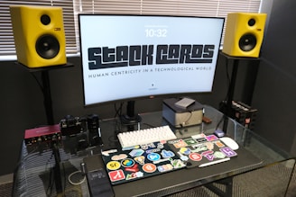 A modern workspace setup featuring a large curved monitor with the words 'STACK CARDS' and 'HUMAN CENTRICITY IN A TECHNOLOGICAL WORLD' displayed. Surrounding the monitor are two vibrant yellow speakers. On the glass desk surface, there is a laptop decorated with numerous colorful stickers, a small computer cube, a keyboard, a smart device, a camera, and some audio equipment. The blinds are partially closed, suggesting a softly lit room.