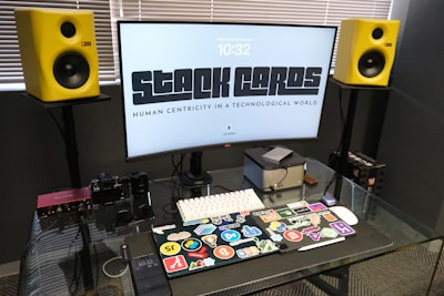 A modern workspace setup featuring a large curved monitor with the words 'STACK CARDS' and 'HUMAN CENTRICITY IN A TECHNOLOGICAL WORLD' displayed. Surrounding the monitor are two vibrant yellow speakers. On the glass desk surface, there is a laptop decorated with numerous colorful stickers, a small computer cube, a keyboard, a smart device, a camera, and some audio equipment. The blinds are partially closed, suggesting a softly lit room.