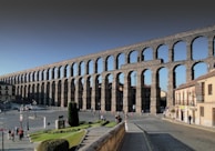 A large, ancient stone aqueduct with multiple arches extends across a cityscape. The structure is prominent against a clear blue sky, with people walking and gathering in the open area below. Surrounding the aqueduct, there are traditional buildings and neatly maintained lawns.