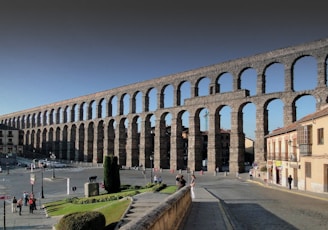 A large, ancient stone aqueduct with multiple arches extends across a cityscape. The structure is prominent against a clear blue sky, with people walking and gathering in the open area below. Surrounding the aqueduct, there are traditional buildings and neatly maintained lawns.